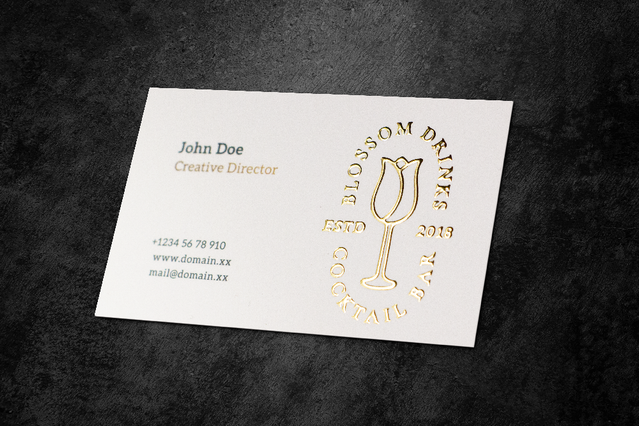 Une carte de visite blanche premium 'Blossom Drive' avec un élégant logo crest tulipe circulaire en dorure à chaud, et les coordonnées de John Doe, Directeur Créatif, sur fond pierre sombre. Les cartes de visite dorure or avec marquage à chaud sont l'outil de communication le plus élégant pour se démarquer.