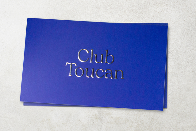 Deux cartes de visite bleu cobalt avec 'Club Toucan' en typographie sérif gauffré argent, empilées sur surface blanche texturée. Les cartes de visite premium avec dorure argent et gaufrage créent une impression tactile luxueuse — idéales pour clubs exclusifs et marques premium cherchant des finitions métalliques sophistiquées.