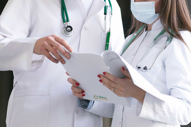 Deux médecins en blouse blanche avec stéthoscopes verts consultant une chemise pour rapports médicaux brandée dans un couloir clinique. Sprint24 imprime des chemises porte-rapports médicaux personnalisées avec découpe et logo pour médecins, hôpitaux et cliniques privées.