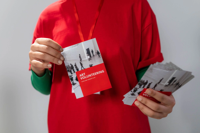 Une personne en chemise rouge tient une pile de cartes postales promotionnelles personnalisées en rouge et blanc pour un événement 'Art Volunteering'. La scène illustre la facilité de distribution des cartes postales lors de foires et événements. Imprimez des cartes postales promotionnelles en ligne avec Sprint24.