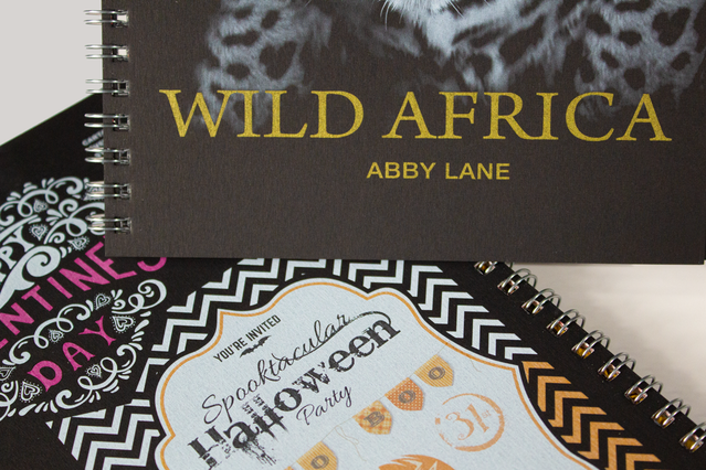 Trois cahiers à spirale présentant des techniques de finition d'impression premium — couverture sombre 'Wild Africa' avec titre doré à chaud et texture animale, design Saint-Valentin avec vernis UV sélectif et invitation Halloween orange. Le catalogue d'échantillons permet d'explorer dorure or, argent coulé et vernis UV.